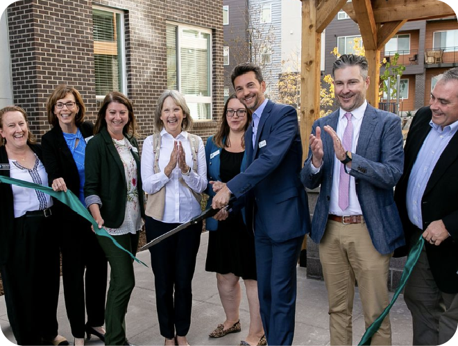 People gathered around, clapping and celebrating, as a man in the middle cuts a forest green ribbon.